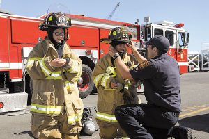 Firefighter Alex Donald helps Raphael and Michael Gravel from Big Brothers and Big Sisters dress in firefighting gear during a tour of Canadian Forces Base Esquimalt July 29. Photo by Cpl Stuart MacNeil, MARPAC Imaging Services