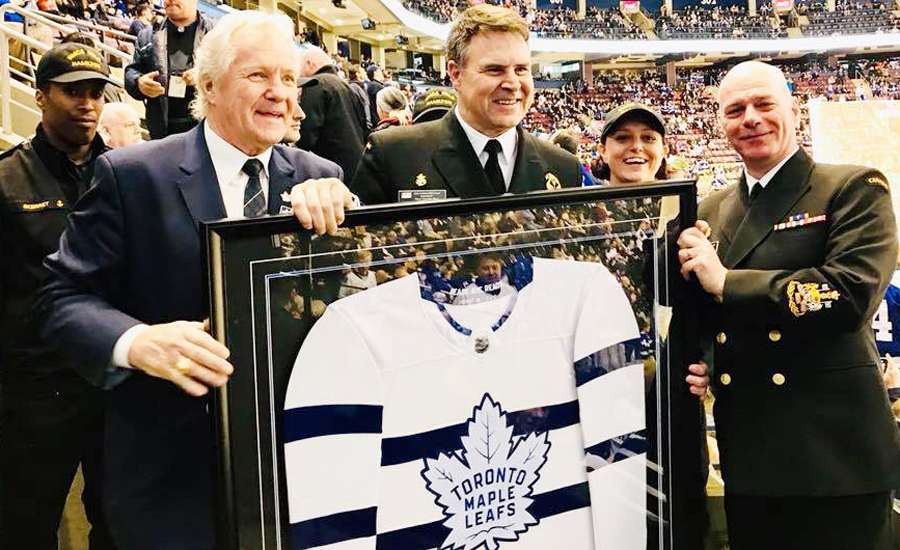 Vice-Admiral Ron Lloyd, centre, Commander of the Royal Canadian Navy, and Command Chief Petty Officer Michel Vigneault, right, accept a framed version of the new RCN Stadium Series Sweater from Toronto Maple Leafs alumnus Darryl Sittler.