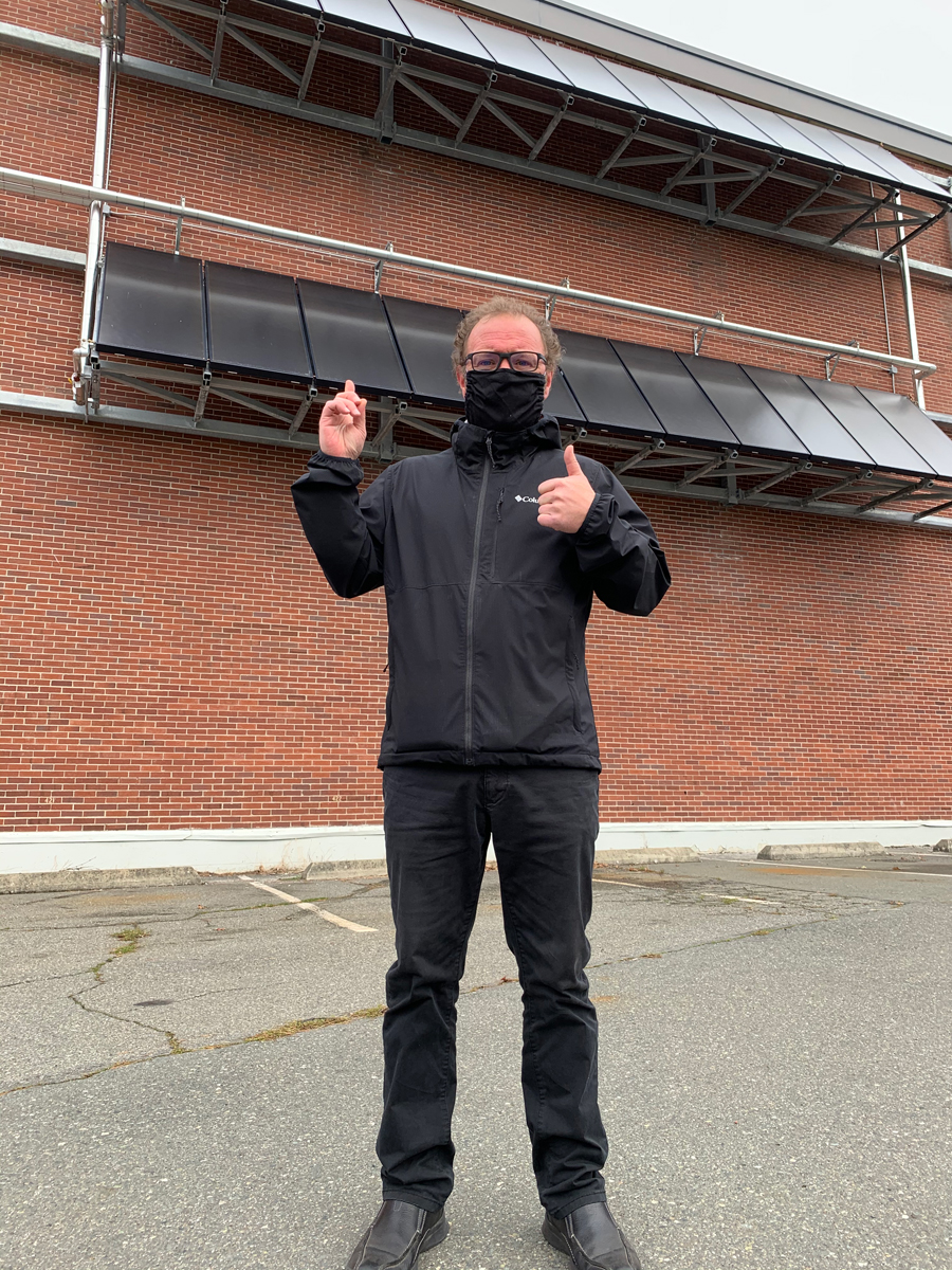 Jamie Birtwistle, Real Property Operations (Esquimalt) Energy Manager, stands below to two rows of solar panels on the west wall of the Naden Athletic Centre. Photo by RPOps (Esq)