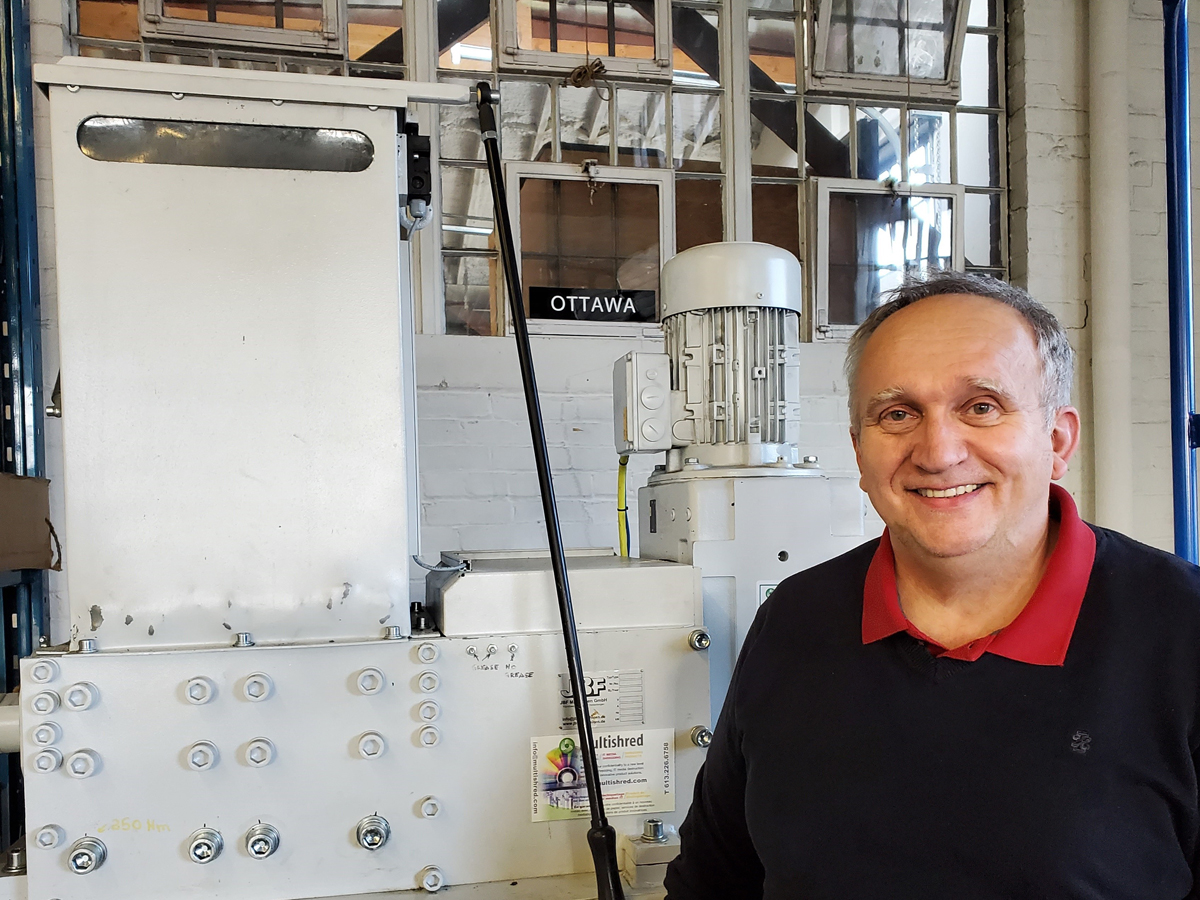 Daniel Bezna of Base Logistics’ Disposal Stores Warehouse, stands in front of a large industrial shredder. The shredder is used to break down some of the approximately 12,000 discarded IT and computer items Bezna handles each year. Photo Credit: BLOG