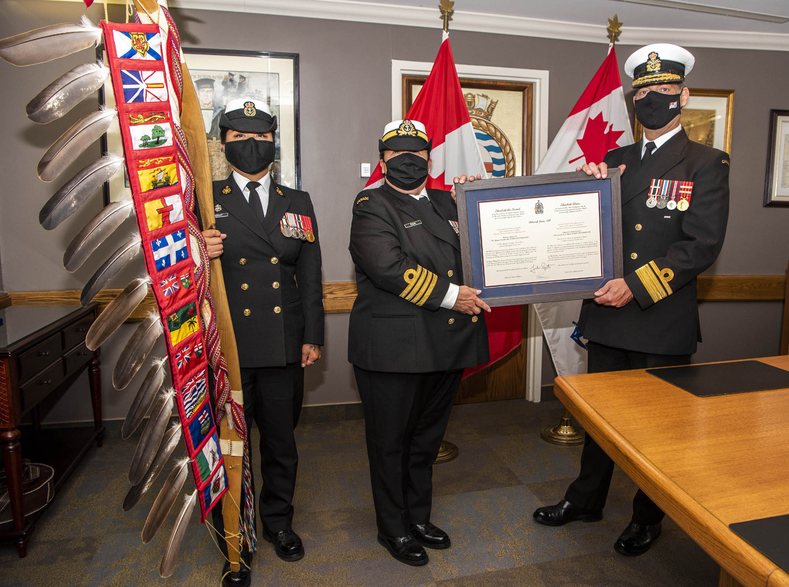 HCapt(N) Debbie Eisan, centre, receives her scroll from RAdm Brian Santarpia at a ceremony on June 21. PO1 Katerina Stewart carries the DND/CAF Eagle Staff.